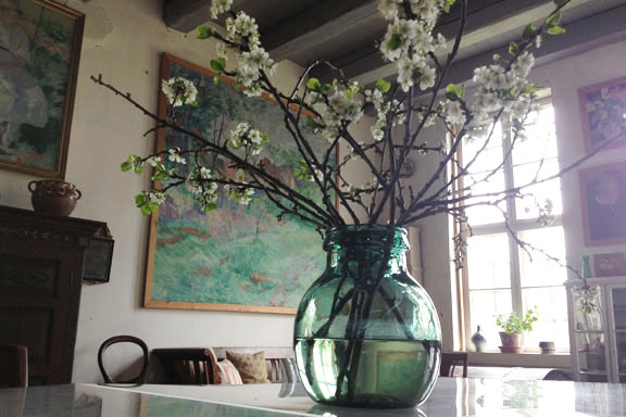 En smuk grønblå vase med friske grene fra haven pryder bordet midt i atelieret. Fotograf: Susanne Randers